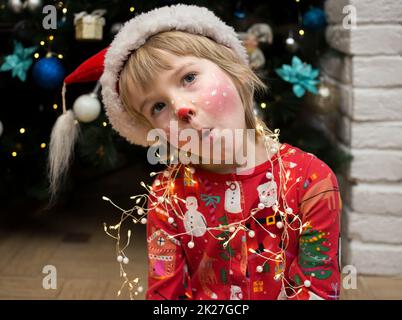 Kind im Weihnachtsschlafanzug, Weihnachtsmütze und ein Hirsch-Make-up auf seinem Gesicht machen lustige Gesichter. Weihnachten, Neujahr, fröhliche Winterferien, Verwöhnung Stockfoto