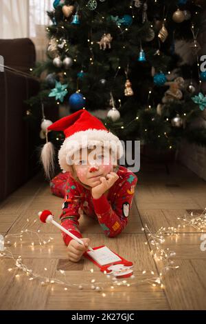 Kind in Weihnachten Pyjamas, Weihnachtsmann Hut und Hirsch Make-up schreibt Brief an den Weihnachtsmann auf dem Boden vor Weihnachtsbaum liegen. Winterferien, inte Stockfoto