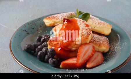 Hüttenkäse-Fritters oder Syrniki oder Pfannkuchen mit frischen Heidelbeeren, Erdbeeren und Erdbeermarmelade. Leckeres Dessert oder süßes Frühstück. Russische und ukrainische Küche Stockfoto