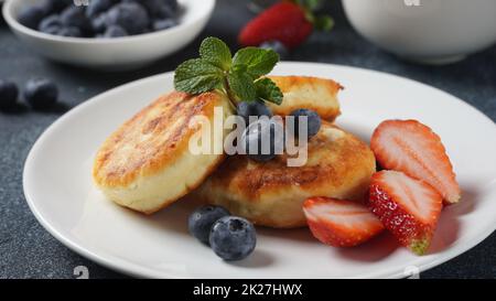 Hüttenkäse-Fritters oder Syrniki oder Pfannkuchen mit frischen Heidelbeeren, Erdbeeren und Erdbeermarmelade. Leckeres Dessert oder süßes Frühstück. Russische und ukrainische Küche Stockfoto