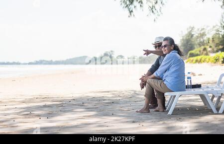 Glückliche asiatische Familie, Seniorenpaar, das auf Liegen mit Rückenlehne sitzt, während eines Strandurlaubs, der sich miteinander unterhält Stockfoto