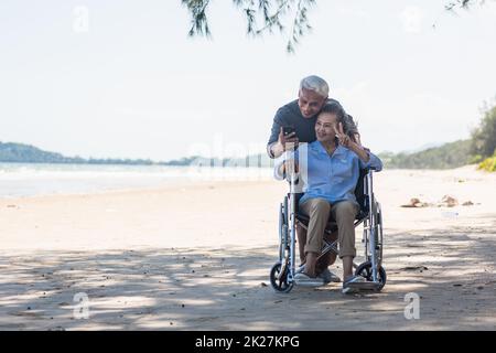 Ältere Frau im Rollstuhl sitzt und Ehemann ist ein Rollstuhl Benutzer Smartphone macht Selfie am Strand Stockfoto