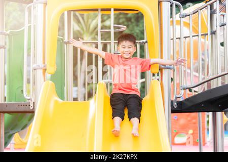 Ein asiatisches Kind lächelt auf dem Spielplatz mit Schiebebügel, ein glückliches Kind im Vorschulalter, das tagsüber im Sommer auf der Spielplatzausrüstung spielt, Klettern eines kleinen Jungen Stockfoto