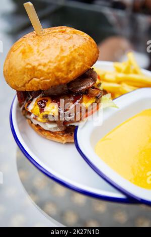 Das Konzept von Fast Food und Essen zum Mitnehmen. Ein saftiger Hamburger liegt neben pommes frites auf einem Metallteller zusammen mit Cheddar Cheese Sauce im Hintergrund. Stockfoto