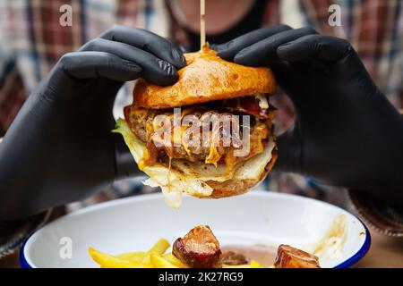 Das Konzept von Fast Food und Essen zum Mitnehmen. Ein Mann in schwarzen Latexhandschuhen hält einen saftigen Hamburger in den Händen, liegt neben pommes Frites auf einem Metallteller zusammen mit Käsesauce. Stockfoto