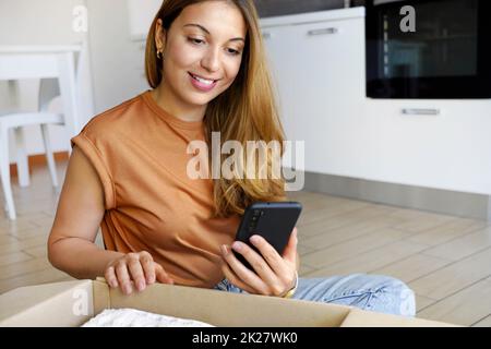 Online-Shop, der Kleidung auf einer Website verkauft, die von zu Hause aus mit einem Smartphone im E-Commerce arbeitet. Frau, die neue Bekleidungskäufe in Versandpaketen zur Lieferung verpackt. Stockfoto