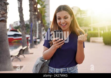 Attraktive junge Frau, die beim Sonnenuntergang auf ihrem Smartphone gute Nachrichten feiert Stockfoto