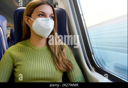 Porträt einer jungen Frau, die eine schützende Gesichtsmaske FFP2 KN95 trägt, mit öffentlichen Verkehrsmitteln, die durch das Fenster blickt Stockfoto