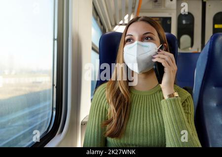 Junge Geschäftsfrau, die eine Schutzmaske trägt, die für öffentliche Verkehrsmittel erforderlich ist, die mit einem Telefon im Zug telefonieren Stockfoto