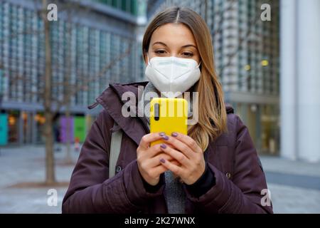 Frontansicht der Winterfrau mit FFP2 KN95 Schutzmaske chatten mit Smartphone in der Stadt Straße Stockfoto