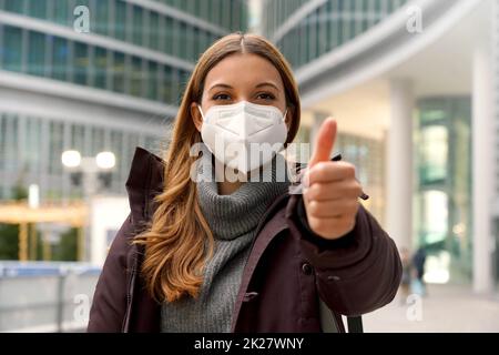 Selbstbewusste Frau trägt Schutzmaske KN95 FFP2 zeigt Daumen nach oben und Blick auf die Kamera in der modernen Stadt Stockfoto