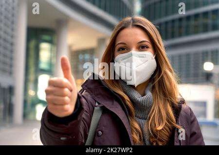 Nahaufnahme einer schönen selbstbewussten Frau, die Schutzmaske KN95 FFP2 trägt und den Daumen mit der modernen Stadt auf dem Hintergrund zeigt und die Kamera anschaut Stockfoto