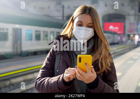 Porträt eines Mädchens in einer Schutzmaske, das auf dem Bahnsteig des Bahnhofs mit einem Handy steht, das das Ticket online kauft Stockfoto