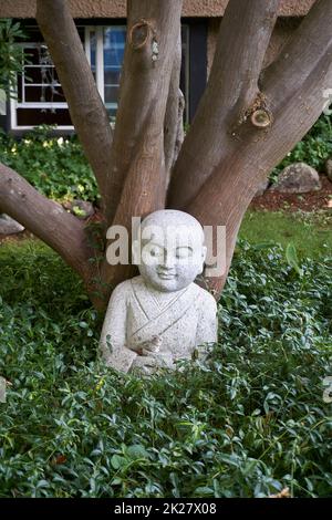 Granitskulptur eines Buddha und eines Hasen, die unter einem Baum inmitten von Grün sitzen Stockfoto