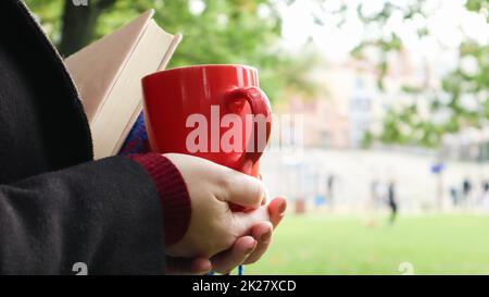 Eine rote Tasse Kaffee und ein Buch mit einer blauen karierten Wolldecke oder in den Händen einer Frau, die im Park einen Pullover und einen schwarzen Mantel trägt. Warmes und sonniges Wetter. Weiche, gemütliche Fotografie Stockfoto