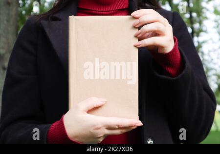 Nahaufnahme einer Frau in schwarzem Wollmantel und burgunderfarbenem Pullover mit einem Buch mit leerem Einband im Park. Freier Platz für das Hintergrundlayout Ihres Buchlesekonzepts. Stockfoto