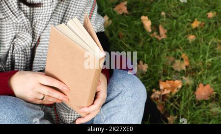 Ein Mädchen liest ein Buch im Herbstpark. Weibliche Hände öffnen die Seiten eines Buches im Freien an einem warmen, sonnigen Tag. Der Schüler bereitet sich auf die Prüfung vor. Literarische Freizeitaktivitäten in der Natur. Stockfoto