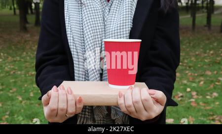 Eine junge Frau in einem schwarzen Mantel und einem grauen Schal hält ein Buch und ein Glas mit Kaffee in den Händen vor dem Hintergrund eines Herbstparks. Im Freien. Bildung, nahe an der Hand des Mädchens. Stockfoto