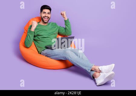 Foto in voller Größe von jungen hispanischen Mann sitzen Sitzsack heben Fäuste Job Angebot gekleidet trendy grünes Kleidungsstück auf violettem Hintergrund isoliert Stockfoto