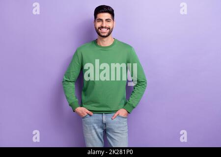 Foto von positiven fröhlichen Kerl gekleidet grün lange Ärmel stehende Hände in den Taschen toothy lächelnd isoliert auf lila Farbe Hintergrund Stockfoto