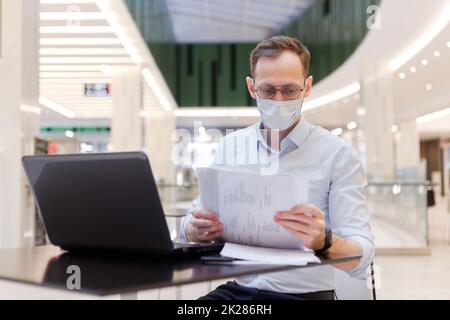 Angestellter mit Gesichtsmaske, Mann, der an einem öffentlichen Ort an einem Laptop arbeitet Stockfoto