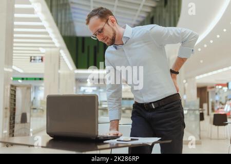 Der Mensch hält sich am Rücken fest, leidet unter Rückenschmerzen, wenn er mit einem Computer neben einem Desktop steht. Sitzender Büroangestellter Stockfoto