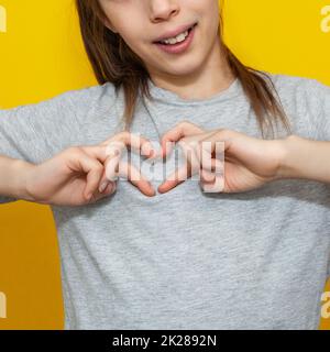 Ein bezauberndes Mädchen im grauen T-Shirt, das eine Herzgeste mit den Fingern macht, mit einem fröhlichen, aufrichtigen Lächeln isoliert auf gelbem Hintergrund. Stockfoto