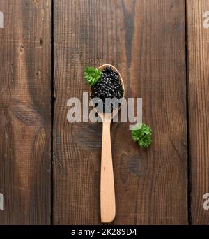 Frischer körniger schwarzer Paddelfisch-Kaviar in braunem Holzlöffel auf schwarzem Hintergrund, Draufsicht Stockfoto