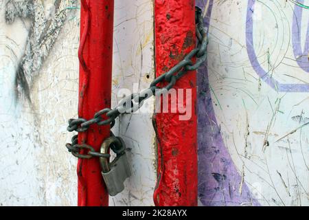 Ein altes Vorhängeschloss auf einer rostigen Kette, die um rote Rohre gewickelt ist. Stockfoto