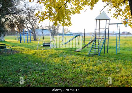 Kinderspielplatz in der Nähe des Stadions vor dem Hintergrund des Sonnenuntergangs. Schaukeln, Rutschen und Leiter. Karussell und Laden. Freizeit für Kinder. Kinderspielplatz. Schaukeln und Rutschen zum Rutschen Stockfoto