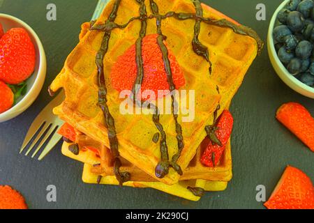 Hausgemachte belgische Waffeln mit Beeren auf Schieferhintergrund. Geschmolzene Schokolade und Schlagsahne auf Waffeln. Flaches Design Stockfoto