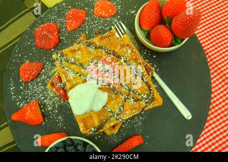 Traditionelle belgische Waffeln mit Heidelbeeren und Erdbeeren. Geschmolzene Schokolade und Schlagsahne auf Waffeln. Draufsicht. Schwarzer Schieferhintergrund Stockfoto