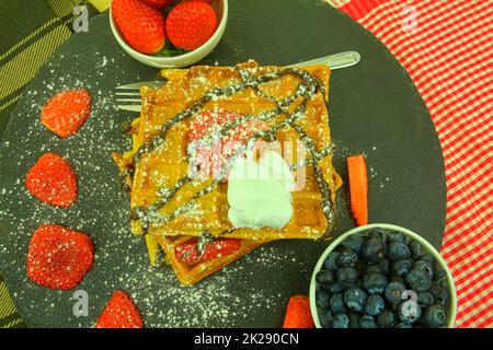 Hausgemachte belgische Waffeln mit Heidelbeeren und Erdbeeren auf Schiefergrund. Geschmolzene Schokolade auf Waffeln. Flaches Design Stockfoto