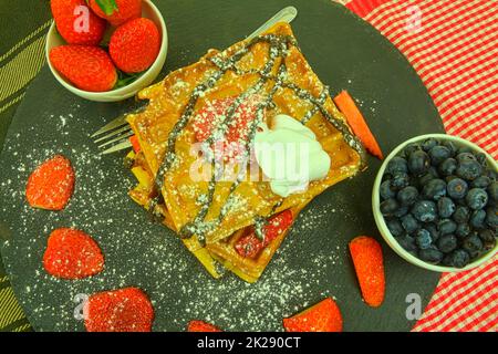 Hausgemachte belgische Waffeln mit Beeren auf Schieferhintergrund. Geschmolzene Schokolade und Schlagsahne auf Waffeln. Flaches Design Stockfoto