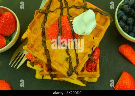 Hausgemachte belgische Waffeln mit Heidelbeeren und Erdbeeren auf Schiefergrund. Geschmolzene Schokolade auf Waffeln. Flaches Design Stockfoto