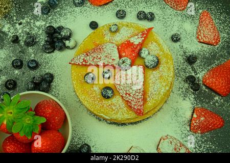 Gesundes Sommerfrühstück, hausgemachte klassische amerikanische Pfannkuchen mit frischen Beeren und Puderzucker. Heidelbeeren und Erdbeeren auf Pfannkuchen. Schwarzer Hintergrund, flach liegend Stockfoto