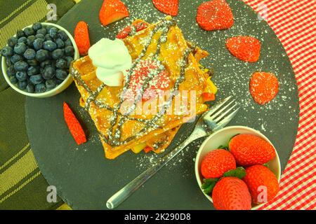 Hausgemachte belgische Waffeln mit Beeren auf Schieferhintergrund. Geschmolzene Schokolade und Schlagsahne auf Waffeln. Flaches Design Stockfoto