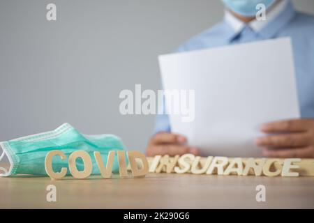 Broker präsentiert über Covid-Versicherungen. Holztext mit grüner medizinischer Maske. Das Objekt befindet sich auf dem Tisch mit Platz an der Wand. Dieser Schuss war Fokus auf den Vordertext. Stockfoto