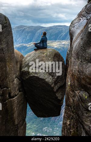 Besuch in Norwegen Kjeragbolten südlich von Lysefjorden die junge Frau sitzt auf dem gian Felsen in meditativer Pose. Stockfoto