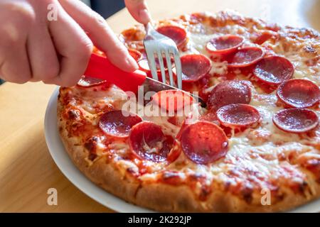 Junge Hände schneiden Salami-Pizza in Stücke mit Gabel und Messer auf Pizzateller in Nahaufnahme Makroansicht mit köstlichem Käse und Salami als fettes, ungesundes Fast Food für hungrige Teenager, italienische Delikatessen Diät Stockfoto