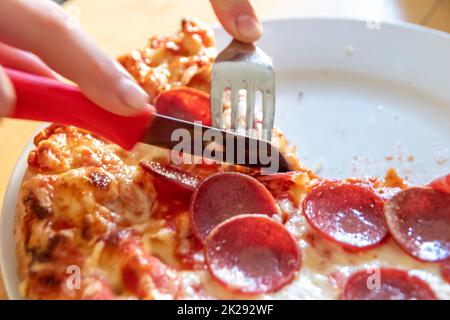 Junge Hände schneiden Salami-Pizza in Stücke mit Gabel und Messer auf Pizzateller in Nahaufnahme Makroansicht mit köstlichem Käse und Salami als fettes, ungesundes Fast Food für hungrige Teenager, italienische Delikatessen Diät Stockfoto