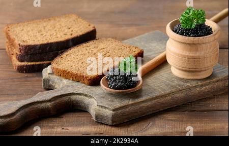 Frischer körniger schwarzer Paddelfisch-Kaviar in braunem Holzlöffel auf einem braunen Tisch, Nahaufnahme Stockfoto