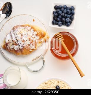 Frühstück am Morgen, rohe Haferflocken auf einem Keramikplatte, Milch in einem Dekanter, Heidelbeeren und Honig in einem Glas auf einem weißen Tisch Stockfoto