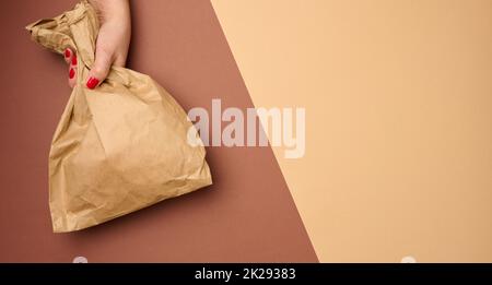 Hand hält einen vollen Papierbeutel aus braunem Kraftpapier auf braunem Hintergrund, Nahaufnahme Stockfoto