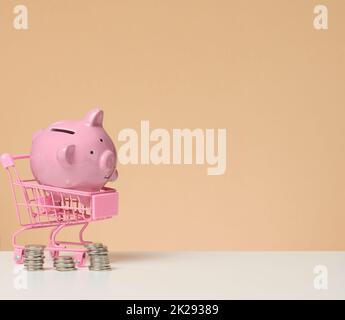 Metall-Mini-Einkaufswagen und rosa Sparschwein auf einem weißen Tisch, brauner Hintergrund. Konzept der Einsparung von Budget, Rabatte Stockfoto