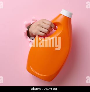 Orangefarbene Plastikflasche mit flüssigem Waschmittel in einer weiblichen Hand auf einem rosa Hintergrund. Ein Teil des Körpers ragt aus einem zerrissenen Loch im Hintergrund heraus Stockfoto