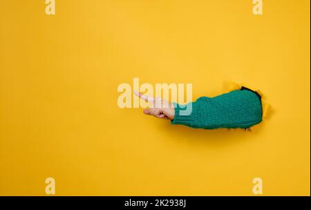 Weibliche Hand ragt aus einem zerrissenen Loch auf einem gelben Papierhintergrund, ein Teil des Körpers zeigt mit dem Zeigefinger zur Seite. Speicherplatz kopieren Stockfoto