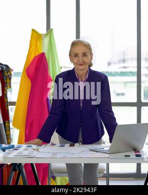 Morgenstimmung im Schneidergeschäft. Senior Designer ist bereit, auf einem Tisch voller bunten Stoffmuster zu arbeiten. Ein Kleiderständer mit bunten Stoffen vor dem großen Fenster. Stockfoto