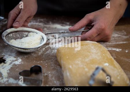 Die Hände des Küchenchefs halten ein Sieb mit Mehl auf dem Hintergrund von Pergament, Mehl, Teig und Keksformen Stockfoto