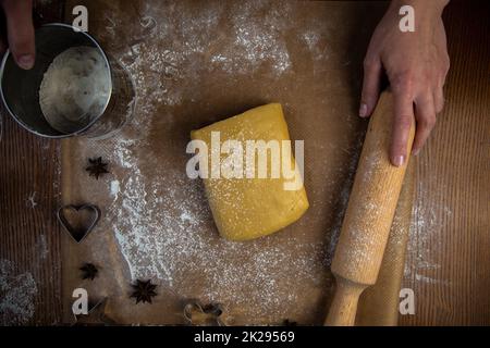 Ein Rechteck aus Teig liegt auf Backpapier, eine Walznadel und Keksschneider liegen daneben, alles ist mit Mehl übersät, ein Koch steht im Hintergrund und hält eine Walznadel und Mehlpulver von oben. Stockfoto